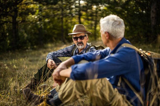 istock Two senior men sitting on grass in forest and talking while taking a break during hiking 1284611389