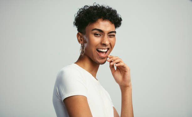 Gender fluid man winking to the camera Gender fluid man with earring winking to the camera. Happy young guy winking  against white background. gender fluid photos stock pictures, royalty-free photos & images