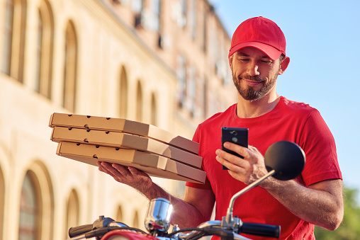 Young cheerful caucasian delivery man in red uniform sitting on scooter and using smartphone while delivering pizza. Food delivery concept