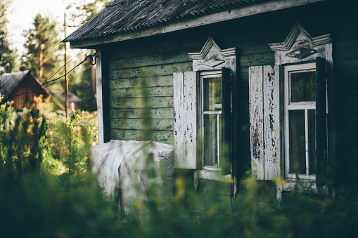 The facade of an old wooden summerhouse in the countryside, in the shadow of the garden with two windows and open shutters, and flaking paint, a slate on a triangular roof, shallow depth of field