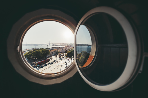 View through a porthole opened round window of an old historic building of a Lisbon landscape with a lively square with tram, bus, and cars, a bridge over the Tagus river in the distance