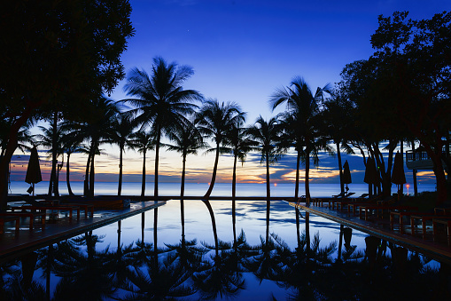 silouette landscape scenery of swimming pool with sunbed and coconut tree at beach front and seaside resort at Huahin Thailand during sunrising