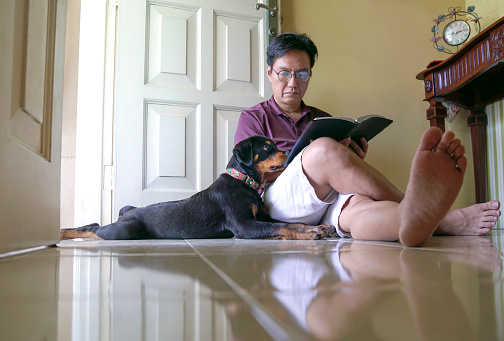 Man reading a book together with pet dog sitting next to him. Indoor setting.