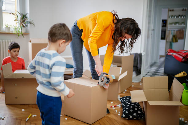 istock Mother bending over and sealing box with tape dispenser, her sons amid boxes 1284675795