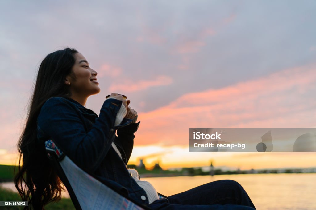 Young woman enjoying hot drink in nature during sunset by lake A young woman is enjoying sitting near a lake and drinking a hot drink in nature during sunset. Women Stock Photo