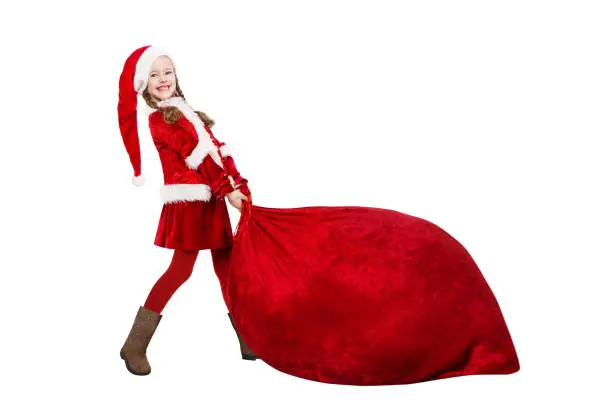 Photo of Little Santa Claus Girl Pulling Huge Red Christmas Bag Full of Gifts.  Funny Child in Santa Hat and Costume Carrying Big Sack with New Year Presents. Isolated White