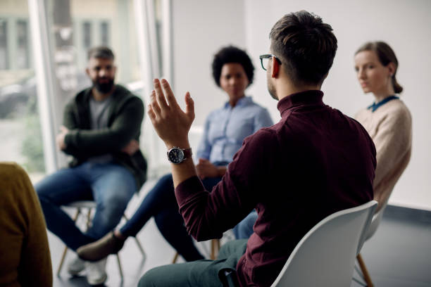 Rear view of man with raised hand on group therapy in medical center. Rear view of man raising his hand to ask a question while attending group therapy. group therapy stock pictures, royalty-free photos & images