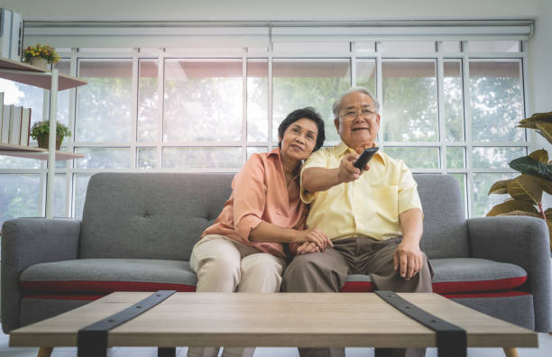 senior asiatische paar lacht und lächelt, während fernsehen zusammen auf einer couch für familien-zweisamkeit. - senior women electronics people remote control stock-fotos und bilder