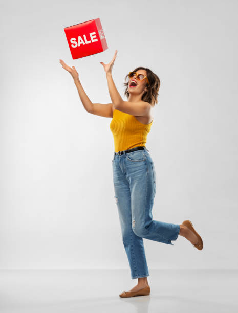 happy smiling young woman posing with sale sign shopping and people concept - happy smiling young woman in sunglasses, mustard yellow top and jeans with sale sign posing over grey background catching stock pictures, royalty-free photos & images