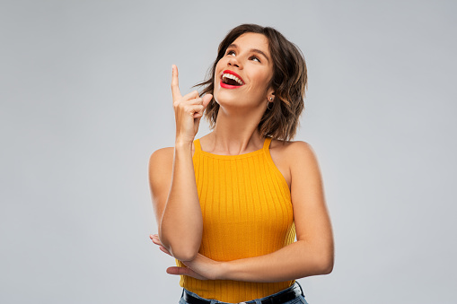 gesture, idea and people concept - happy smiling young woman in mustard yellow top pointing finger up over grey background