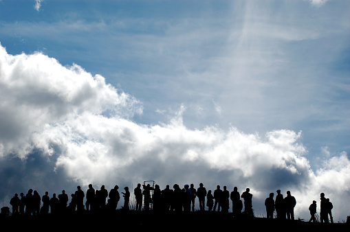 People crowd standing on hill, in front of a cloudy sky.