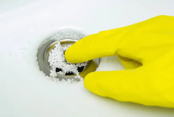 The drain cleaner granules in sink. Hand wearing yellow rubber glove cleaning the plughole of a bathroom sink to unblock the drain