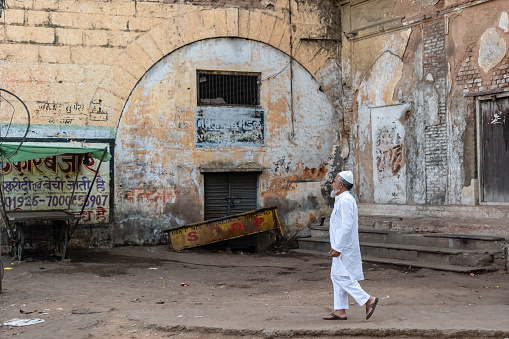 Bhopal, Madhya Pradesh, India - March 2019:An elderly muslim man dressed in a white kurta and a skullcap walking past an old, dilapidated heritage  building in the city of Bhopal.