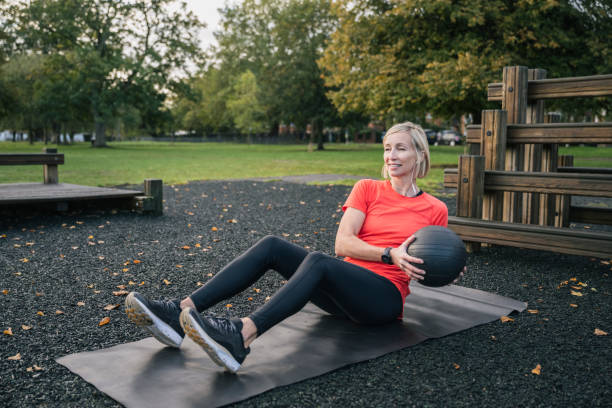 Sportswoman doing seated oblique twist with medicine ball Blonde female athlete in early 50s sitting on exercise mat at public park fitness site working out with medicine ball and strengthening her core. wandsworth photos stock pictures, royalty-free photos & images