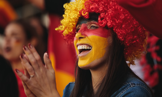 Woman sitting in stadium wearing a wig and her face painted in german flag colors applauding their team. Female from Germany in fan zone watching a soccer match.
