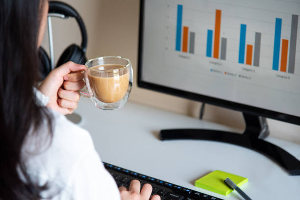 Woman having a cup of coffee while working and using computer on a white desk at home office closeup