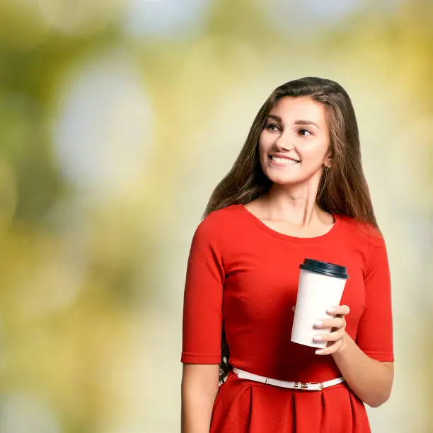 young Girl hold takeaway cup fo coffee. Red dress. White glass with black. Green blur background.