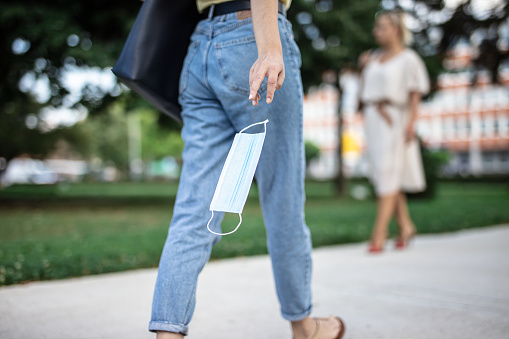 Low section and rear view shot of a woman in jeans walking in a city park and holding a protective face mask in her hand