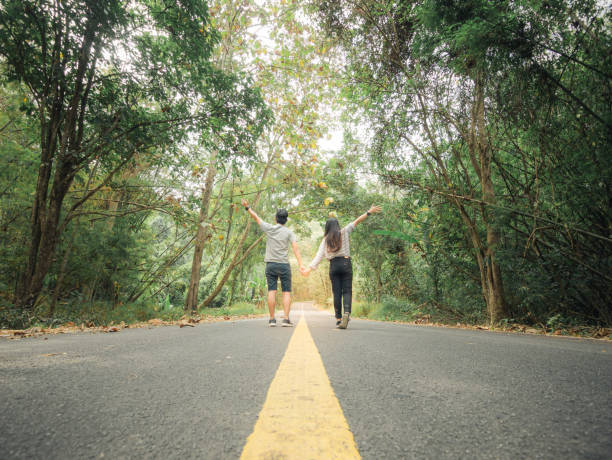 旅行者のカップルは、緑豊かな木々の中で道路を歩いて手をつないでいます。旅行中に自由な時間を楽しんで幸せなカップル。 - two lane highway 写真 ストックフォトと画像