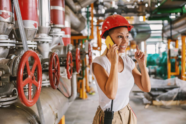 istock Smiling friendly female worker in working suit with protective helmet on head standing next to pipes in heating plant and talking over smart phone with her boss. She is having idea how to solve problem 1284749680