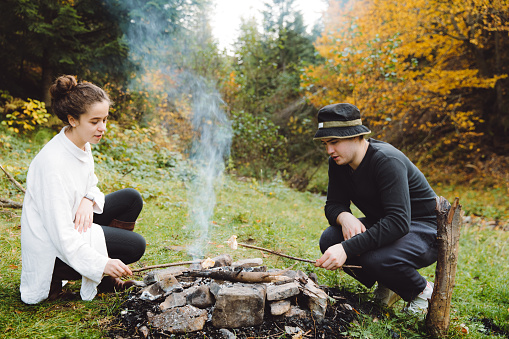 Young couple celebrating Halloween during autumn time and having picnic in the wilderness area on the beautiful meadow near the river , making barbecue at the campfire, talk g and laughing, feeling happiness