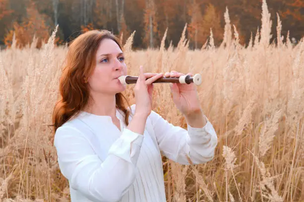 Red hair woman with blockflute play music in autumn field, yellow grass. A musician with a wind instrument in the tall grass near the forest
