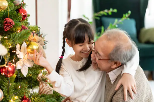 Little girl decorating a Christmas tree with her grandfather. They decorating the Christmas tree prepare for season greeting of Merry Christmas. Multigenerational asian Family concept.
