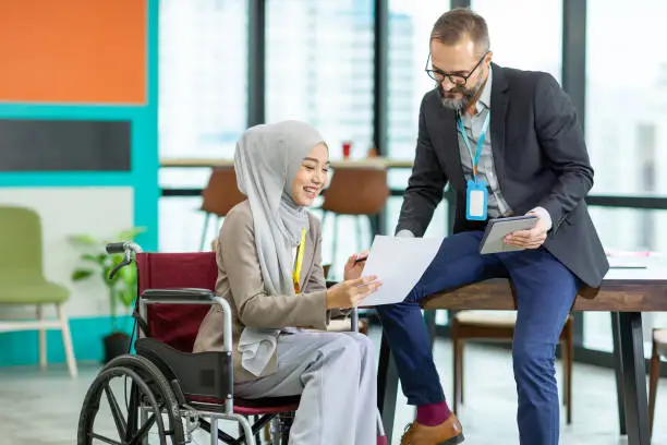 Asian Muslim businesswoman sitting on wheelchair presenting business graph on paper to manager. Smiling group of diverse corporate colleagues in the modern office. Diversity or multicultural in office