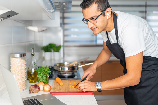 Smiling young man is working with laptop computer  while cooking healthy meal in the kitchen.
