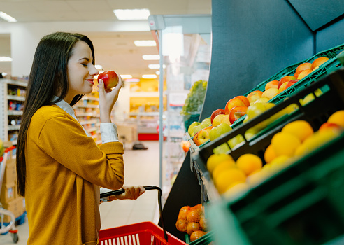 Young woman sniffing apple while doing grocery shopping