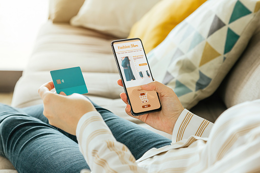 Stock photo of a woman buying a dress online with the phone and a credit card from the sofa at home. E-commerce concept