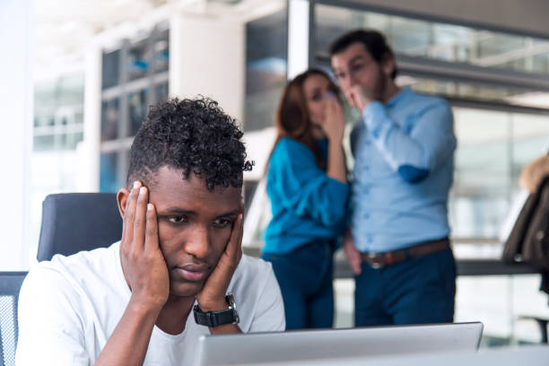 Gossip concept in a modern office A Sad looking Black man is working on desk and there are two colleagues are gossiping at background behind of him aggression stock pictures, royalty-free photos & images