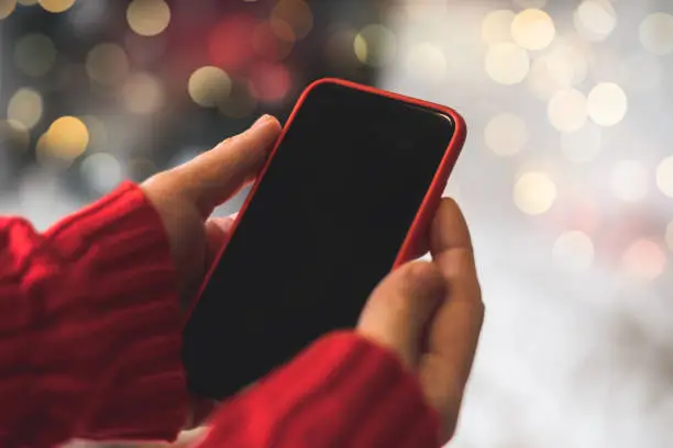 Photo of Cropped of woman's hands in red sweater holding smartphone with blank screen of gadget at Christmas time. Digital mobile phone with copy space area