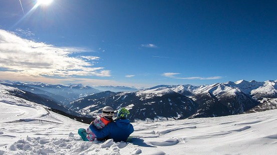 A couple sitting on the snow on top of Katschberg in Austria. They are happy and joyful. Panoramic view on the surrounding mountains. Winter wonderland. Bright and sunny winter day. Love and happiness