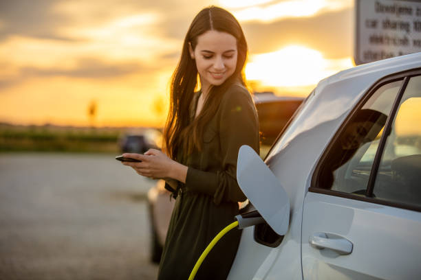 Woman waiting for electric car to charge in the parking lot at sunset Side view waist up shot of a young woman in a dress using her mobile phone while waiting for her white electric car to charge during sunset motor living stock pictures, royalty-free photos & images