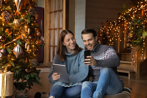 Smiling millennial man and woman sit near decorated Christmas tree use modern gadgets together. Happy young Caucasian couple enjoy leisure winter holidays, have fun browsing devices at home.