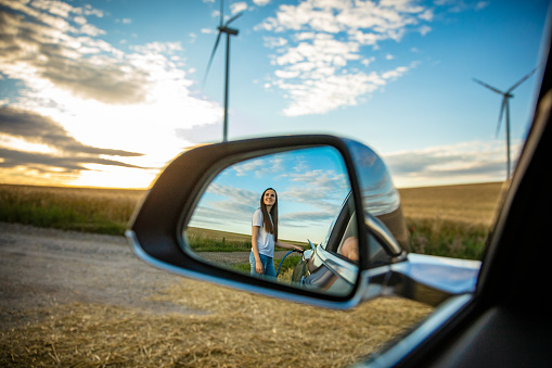 Reflection of young woman charging electric car on road in rural scenery with wind turbines at sunset