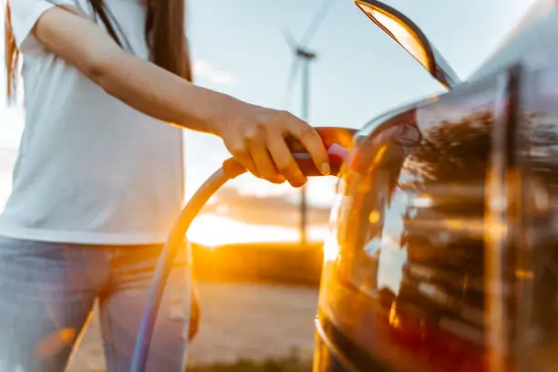 Woman charging electric car on road in rural scenery with wind turbines at sunrise