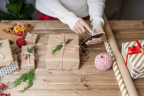 frau hände wickeln weihnachtsgeschenk, nahaufnahme und fotografieren am telefon. unvorbereitete geschenke auf holzhintergrund mit dekorelementen und gegenständen, ansicht von oben. neujahr diy verpackung konzept. - christmas decoration photography themes christmas ornament stock-fotos und bilder