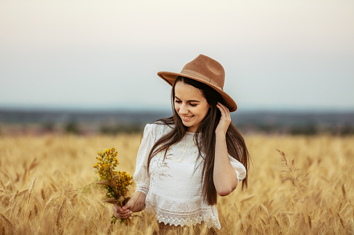 Waist up shot of a smiling young woman with black hair and with yellow flowers in her hand standing in a vast wheat field