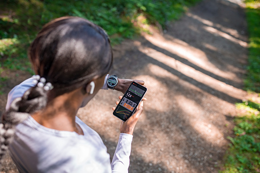 Rear view of woman synchronizing her smart watch and smartphone while checking her heart beats in forest.