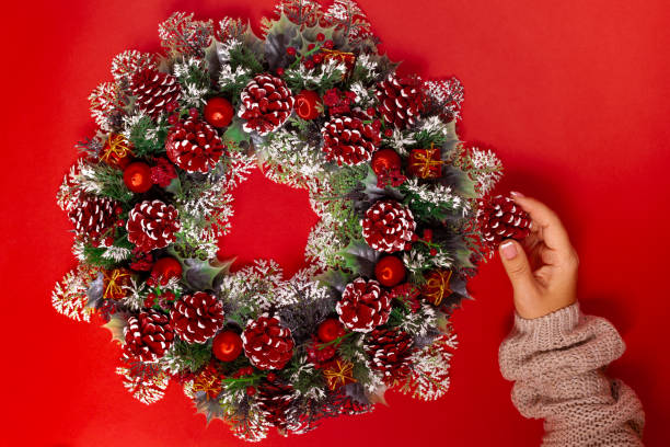 a woman's hand in a beige sweater makes a Christmas wreath of cones ,berries and balls, isolated on a red background a woman's hand in a beige sweater makes a Christmas wreath of cones ,berries and balls, isolated on a red background. Diy Santa Claus Ball Wreath stock pictures, royalty-free photos & images