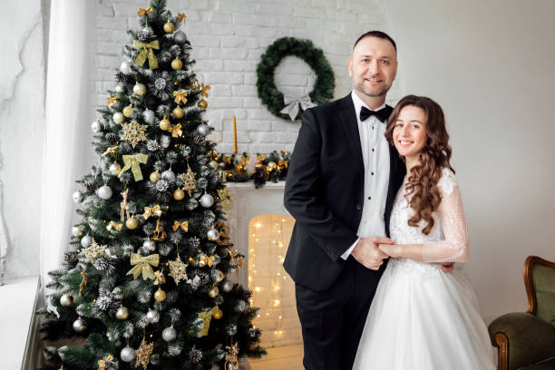 jovem casal apaixonado de noiva e noivo posando em estúdio ao fundo decorado com árvore de natal no dia do casamento no natal. aproveite um momento de felicidade e amor. - christmas wedding bride winter - fotografias e filmes do acervo