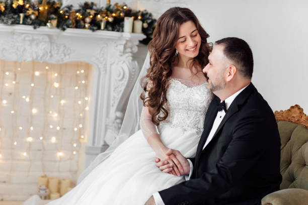 jovem casal apaixonado de noiva e noivo posando em estúdio ao fundo decorado com árvore de natal no dia do casamento no natal. aproveite um momento de felicidade e amor. - christmas wedding bride winter - fotografias e filmes do acervo