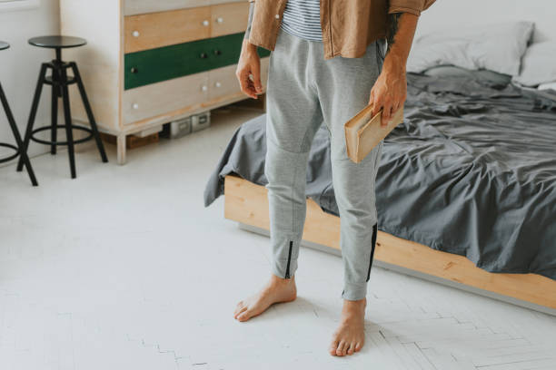 Legs of a Man Standing in the Bedroom with a Book in His Hand - a Close Up Barefoot man reading a book in his bedroom jogging pants stock pictures, royalty-free photos & images