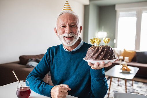 Senior man is at home, he is wearing a party hat and holding a birthday cake