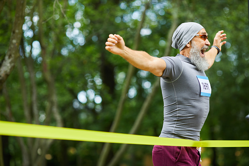 Happy bearded senior man wearing cap and sunglasses winning first place in marathon race, copy space