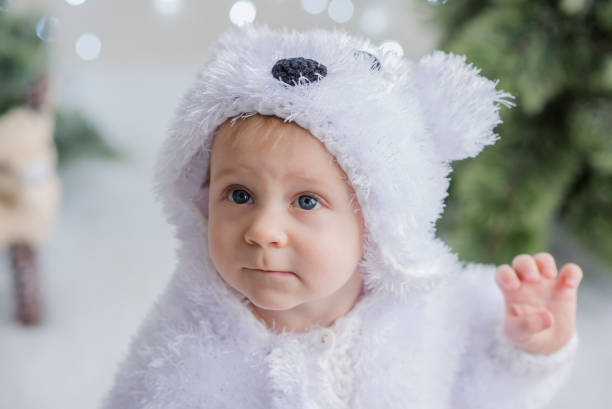 ein kleiner junge, der als weißer eisbär verkleidet ist, sitzt zwischen dem wald der weihnachtsbäume auf einem holzschlitten. in der nähe befindet sich ein redes. verschwommenbokeh lichter hintergrund. neujahrskarte. nahaufnahme porträt - polar bear bear white close up stock-fotos und bilder