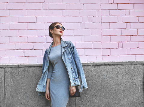 Estilo de calle Shoot mujer en la pared rosa. Swag chica con chaqueta de jeans, vestido gris, gafas de sol. Estilo de vida de moda al aire libre photo