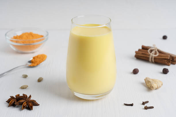 leche dorada servida en vaso alto con varias espigas y cuchara de cúrcuma sobre mesa de madera blanca - turmeric indian cuisine ground white fotografías e imágenes de stock